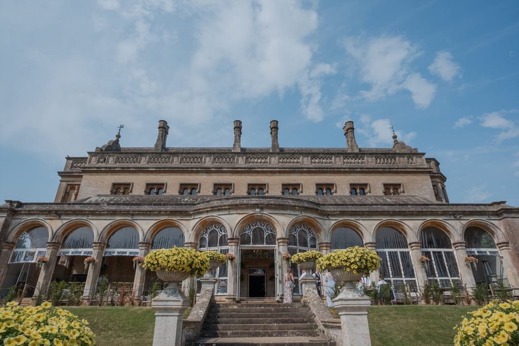 A large, historical building featuring arched windows and multiple chimneys, surrounded by yellow flowers in planters, under a partly cloudy sky—perfect for any Grittleton House photographer to capture its timeless beauty.