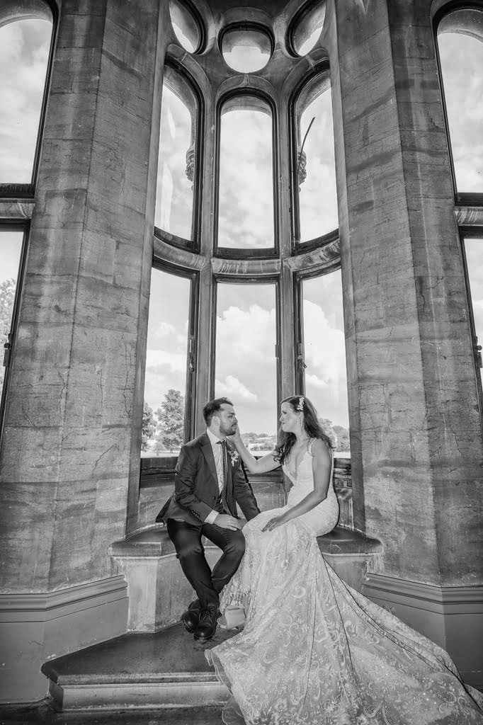 A couple in formal attire sits on a window ledge inside the grand stone building of Grittleton House, captured beautifully by the photographer through its large windows.