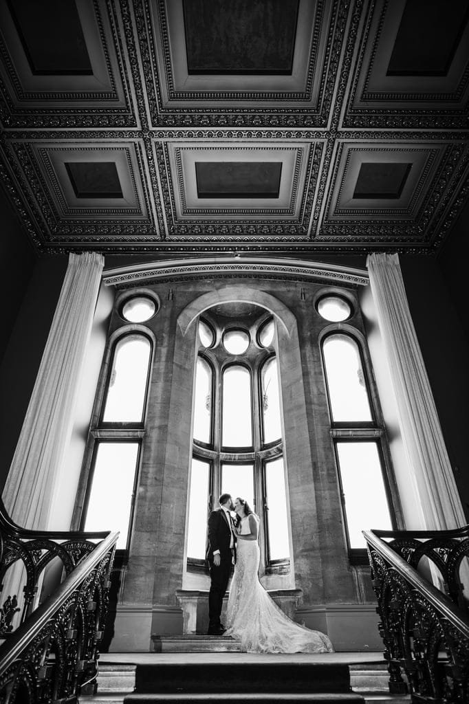 A couple in wedding attire share a kiss by a large, ornate window in an elaborately designed room with intricate ceiling details and grand staircases on either side, captured perfectly by a Grittleton House photographer.