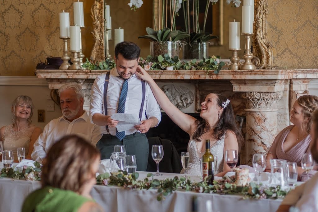 A Grittleton House photographer captures a man reading from a piece of paper while smiling, as a woman beside him touches his arm. They are seated at a table with several others, surrounded by tableware and greenery. The setting appears festive.