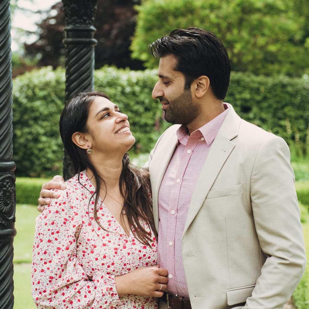 A man and woman stand close together outdoors, smiling at each other. The woman is wearing a white floral dress, and the man is in a beige blazer with a pink shirt. Greenery is in the background, capturing a moment perfect for a Farleigh House Wedding Photographer.