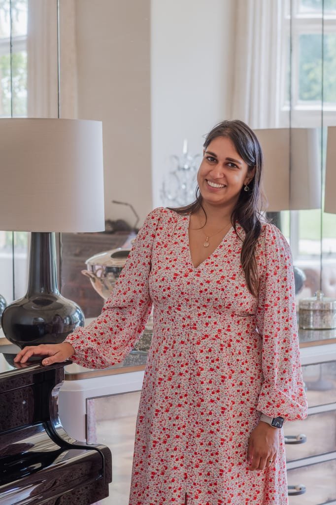 Woman in a floral dress stands smiling next to a piano in a room with large windows and two lamps, capturing the elegance akin to a Farleigh House Wedding Photographer's perfect shot.