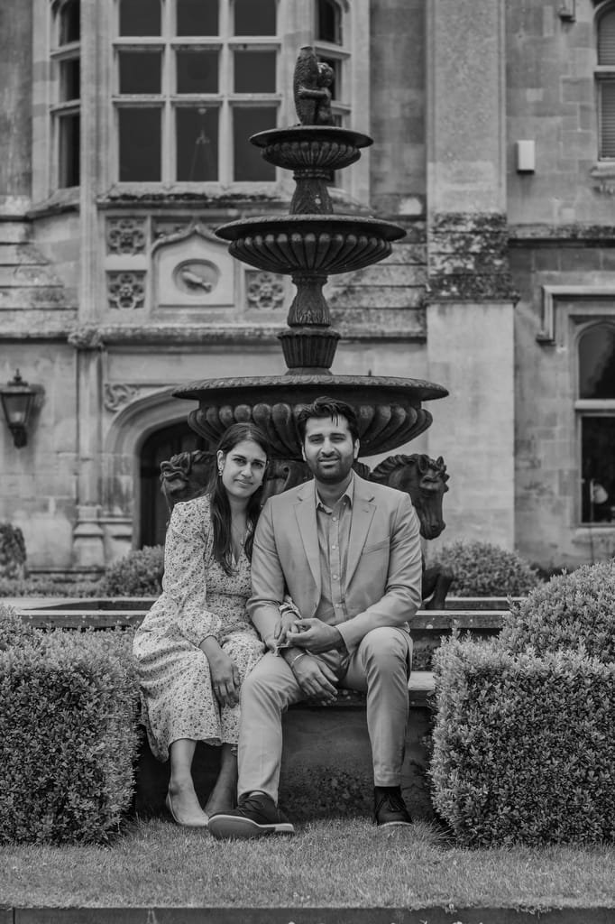 A man and woman sit together in front of a large stone fountain outside a historic building, captured beautifully by a Farleigh House Wedding Photographer. The man is wearing a suit, while the woman is dressed in a patterned dress. The photo is in black and white.