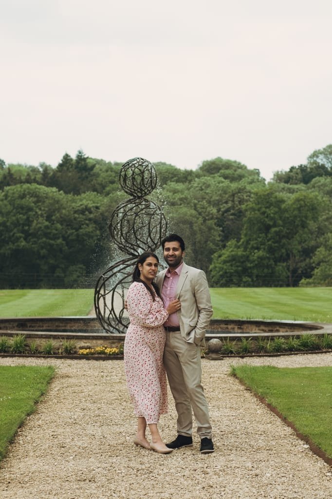 A man and woman stand together on a gravel path in front of a modern outdoor sculpture, with a lush green landscape in the background, evoking the charm often captured by Farleigh House Wedding Photographers.