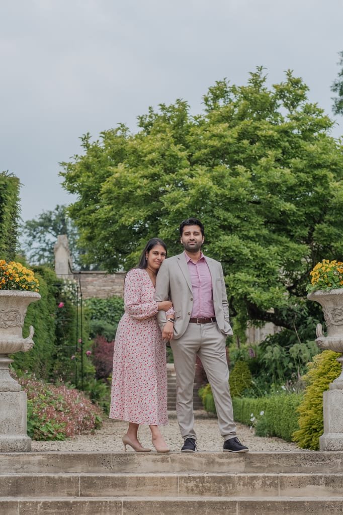 A couple stands on stone steps at Farleigh House, with green trees and plants in the background. The woman wears a floral dress and the man wears a light-colored suit and pink shirt. They appear relaxed, capturing the perfect moment for a Farleigh House Wedding Photographer.