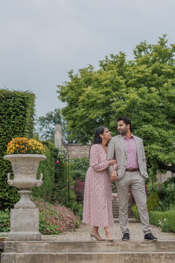 A couple stands on stone steps in a garden, holding hands and smiling at each other. The woman wears a floral dress, and the man is in a light grey suit. A large planter with flowers is in the foreground, perfectly capturing the romantic moment for their Farleigh House Wedding Photographer.