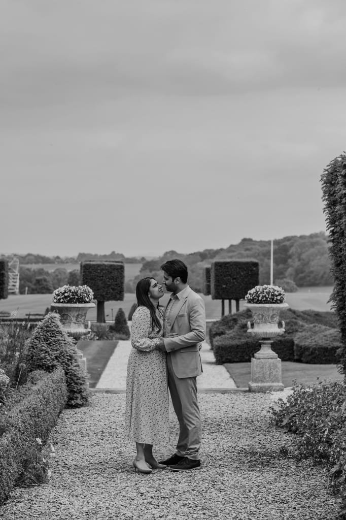 A couple stands close, facing each other and holding hands in a garden pathway lined with topiary and an expansive landscape in the background, captured beautifully by a Farleigh House Wedding Photographer.