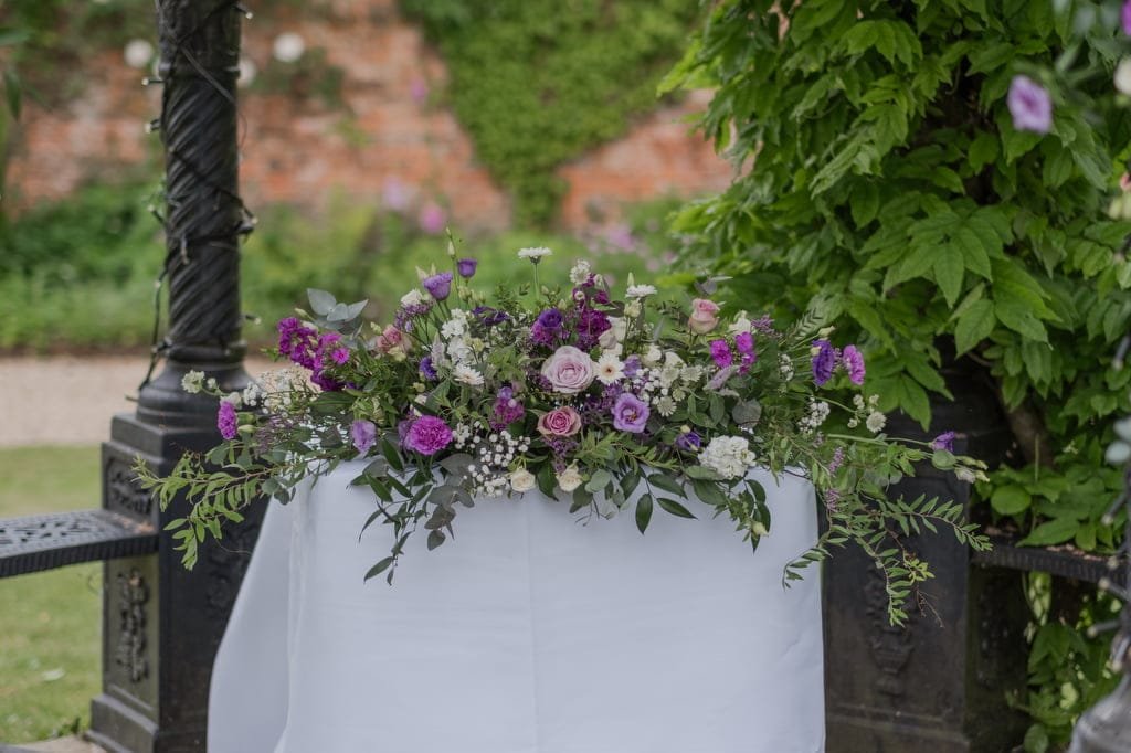 A white table adorned with a floral arrangement featuring purple, white, and pink flowers set in the picturesque outdoor garden of Elmhay Park—a perfect backdrop for weddings captured by a skilled photographer.