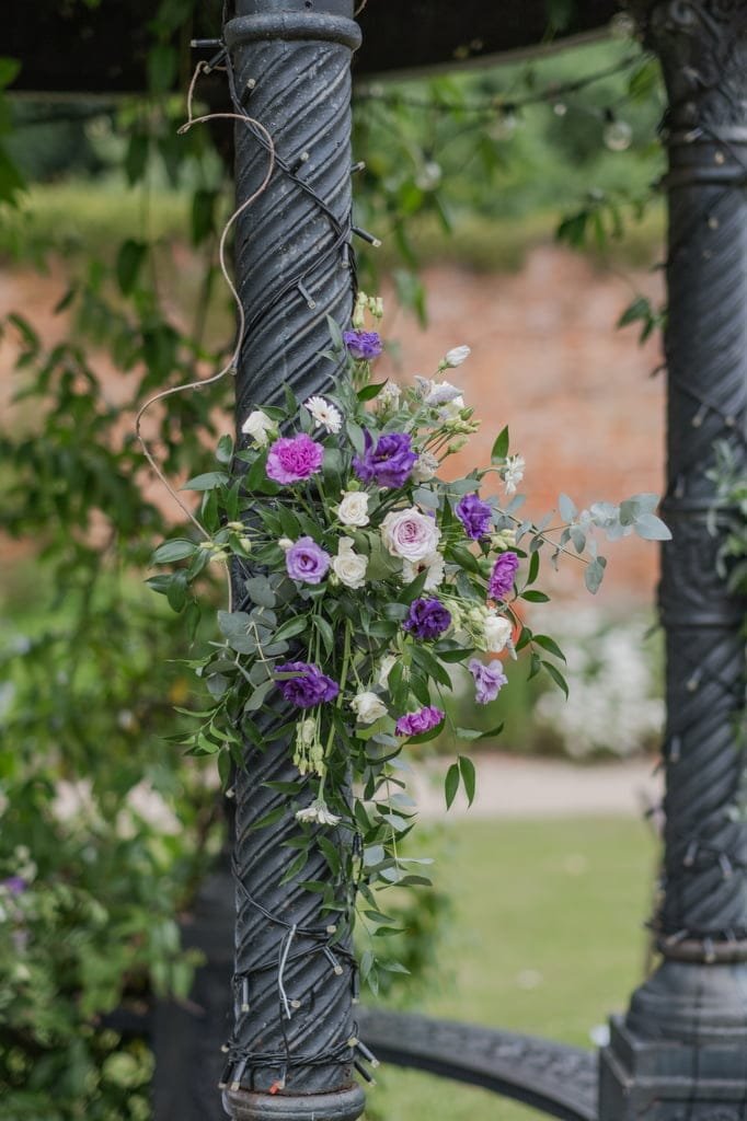 A stunning iron column wrapped with a garland of green leaves and white, purple, and pink flowers stands outdoors, creating a perfect backdrop for weddings at Elmhay Park. The blurred garden background adds a touch of magic that any photographer would love to capture.