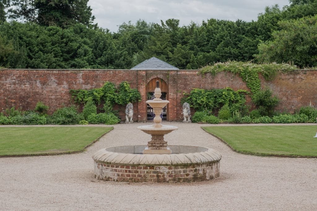 A circular fountain with tiered bowls graces the center of a gravel path at Elmhay Park, surrounded by lush grass and framed by a tall brick wall adorned with greenery and a charming wooden door—a picturesque spot perfect for any weddings photographer.