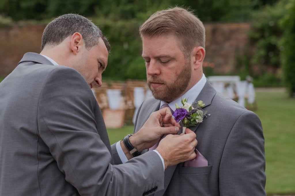 Two men in suits; one man adjusts the other man's boutonniere. The setting is outdoors with greenery and seating in the background, perfectly captured by a skilled weddings photographer at Elmhay Park.