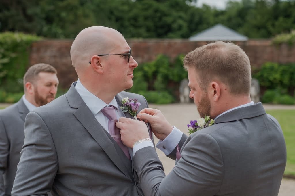 A man adjusts another man's tie and boutonniere while wearing gray suits, standing outside with greenery and a brick wall in the background. Nearby, another man in a gray suit stands by, all captured perfectly by the weddings photographer at Elmhay Park.