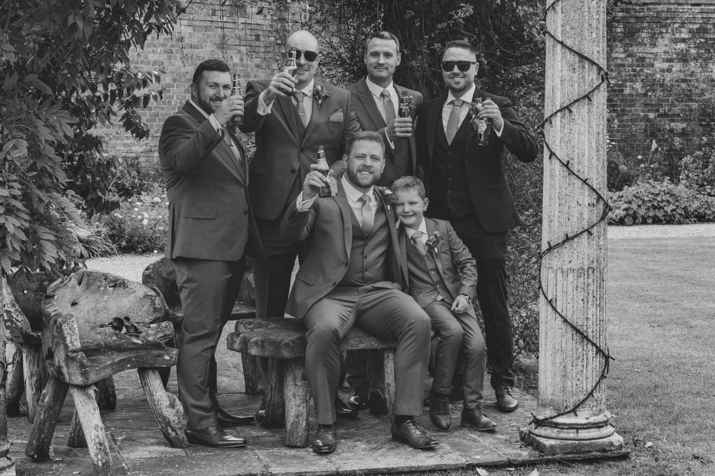 A group of six men, dressed in formal suits, pose together outdoors at Elmhay Park, with some raising drinks in celebration. One man sits on a wooden bench with a child beside him. They are near a column and foliage, perfectly captured by the weddings photographer.