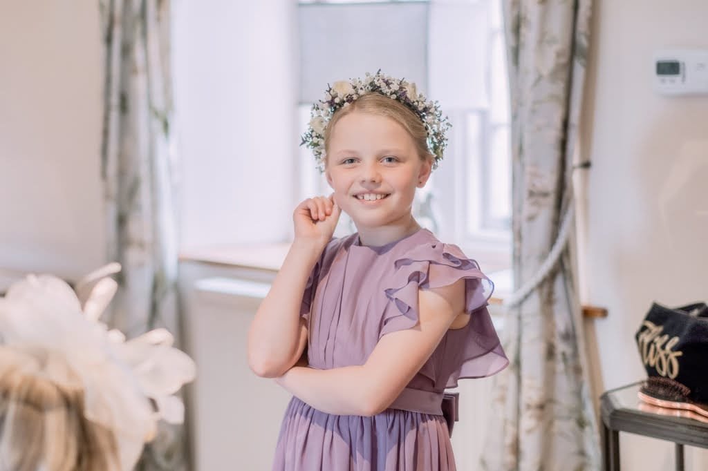 A young girl wearing a lavender dress and floral headpiece stands indoors with her arm crossed, smiling at the camera, perfectly capturing the essence of joyful moments that a talented weddings photographer at Elmhay Park would cherish.