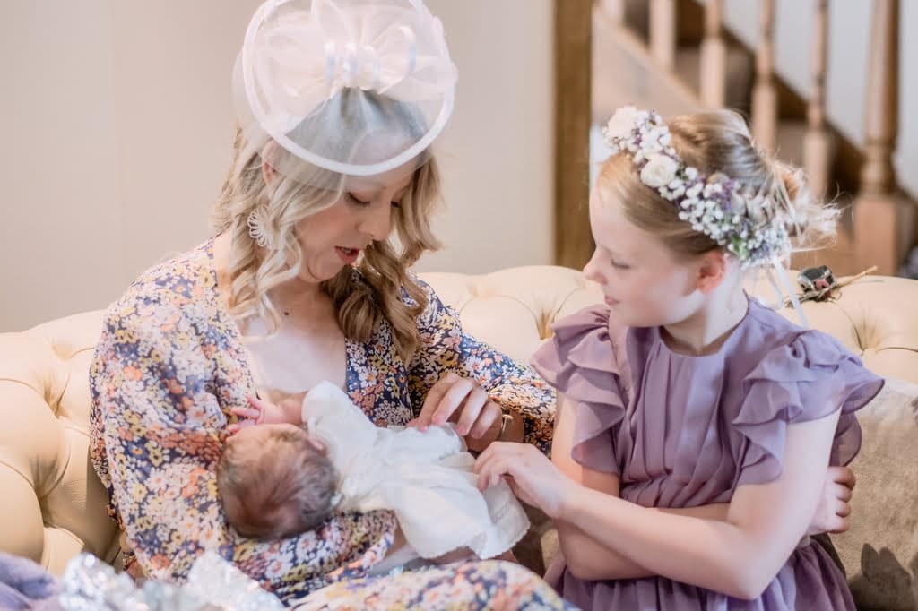 A woman in a floral dress is looking down and holding a baby, while a young girl in a purple dress and flower crown stands beside them on a couch, captured beautifully by the weddings photographer at Elmhay Park.