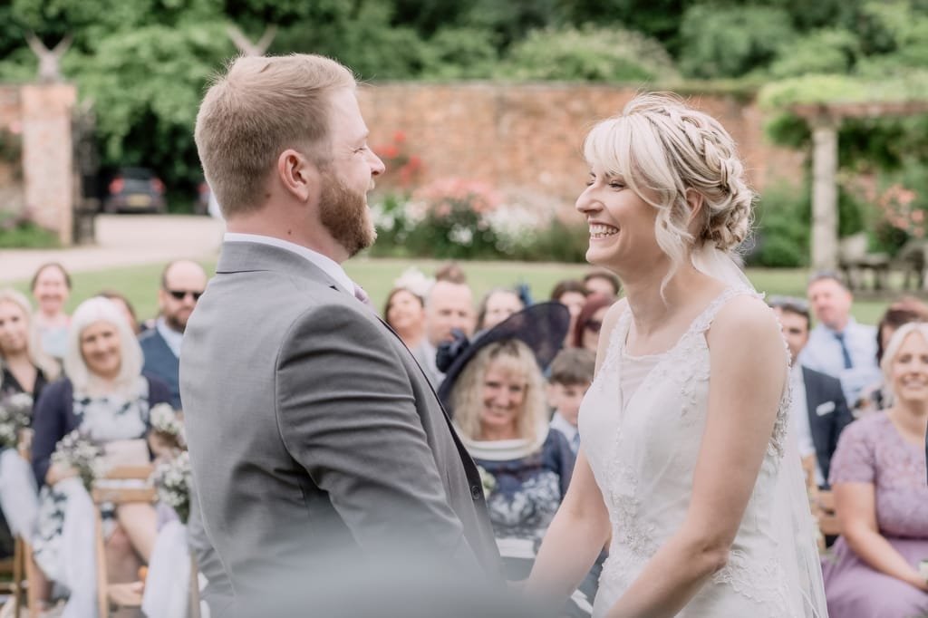 A bride and groom smile at each other during an outdoor wedding ceremony at Elmhay Park, with guests seated in the background, beautifully captured by a weddings photographer.