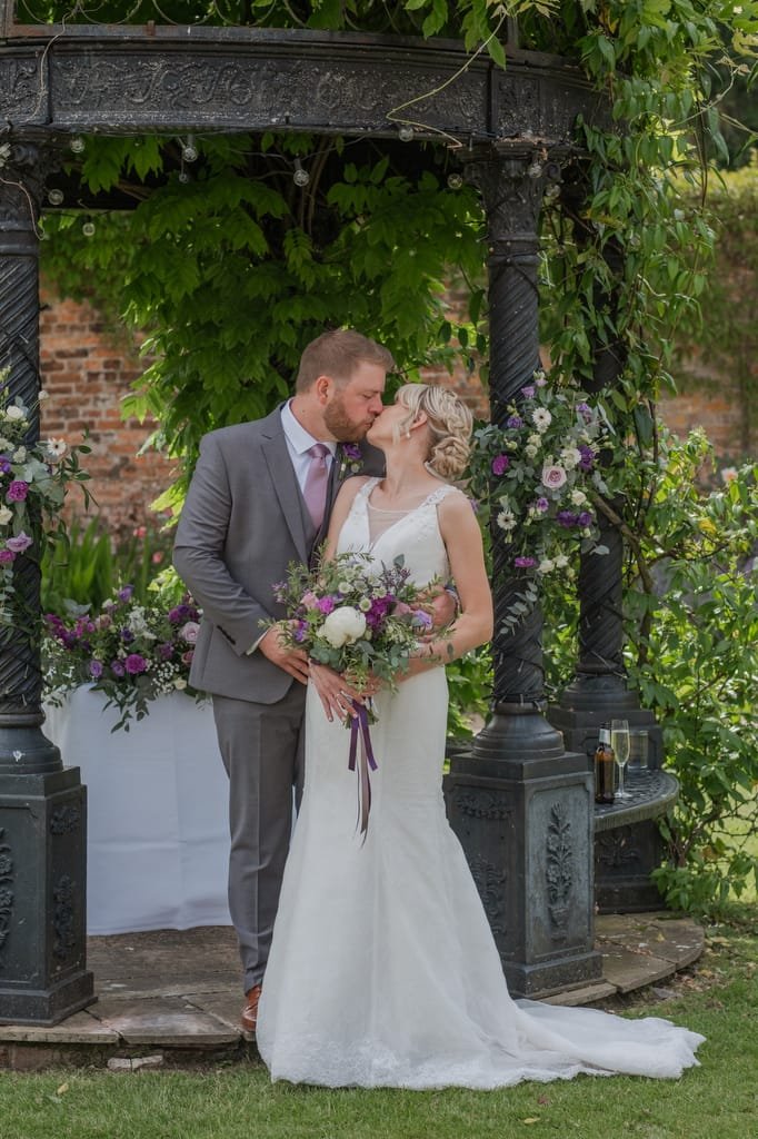 A bride in a white dress holds a bouquet while a groom in a gray suit kisses her on the forehead. They stand under a flower-decorated gazebo, captured beautifully by the weddings photographer at Elmhay Park.