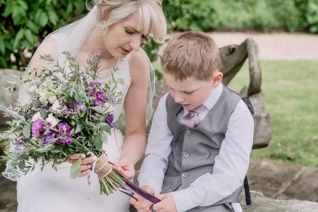 A bride and a young boy, both dressed formally, sit on a wooden bench outdoors at Elmhay Park. The bride holds a bouquet and shows something to the boy. Captured by a skilled weddings photographer, the background is lush with greenery.