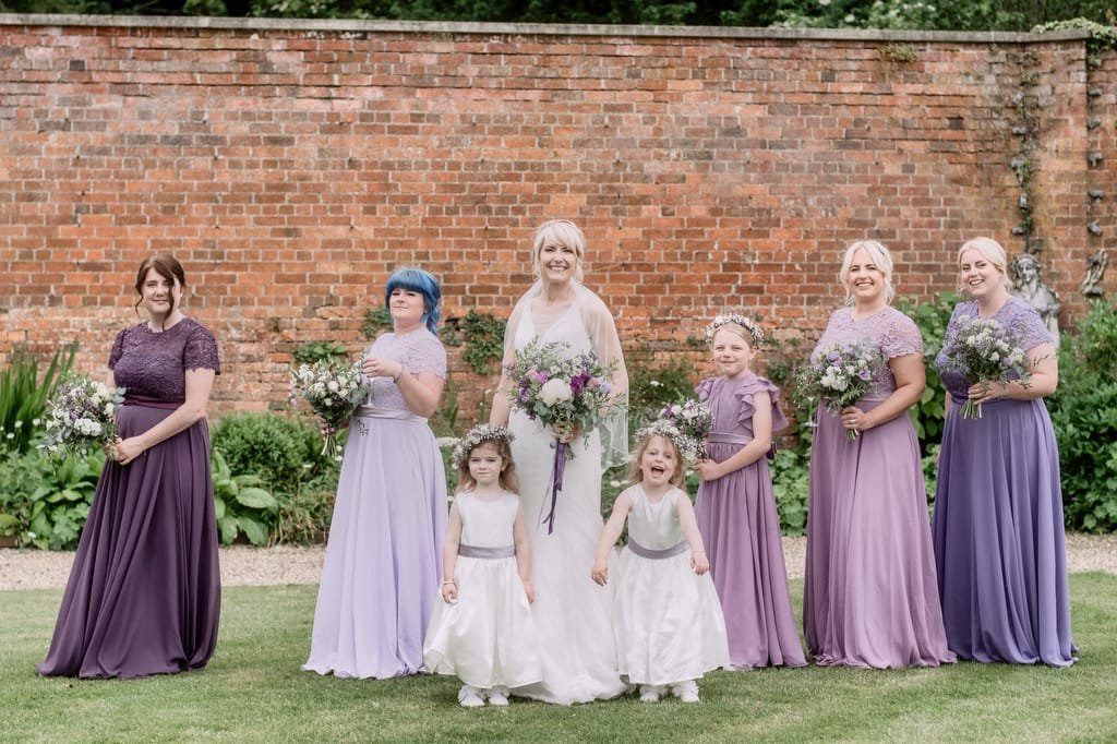 A bride stands with six bridesmaids in lavender and purple dresses, along with two flower girls in white dresses, in front of a brick wall. Captured by a skilled weddings photographer at Elmhay Park, this moment embodies elegance and charm.