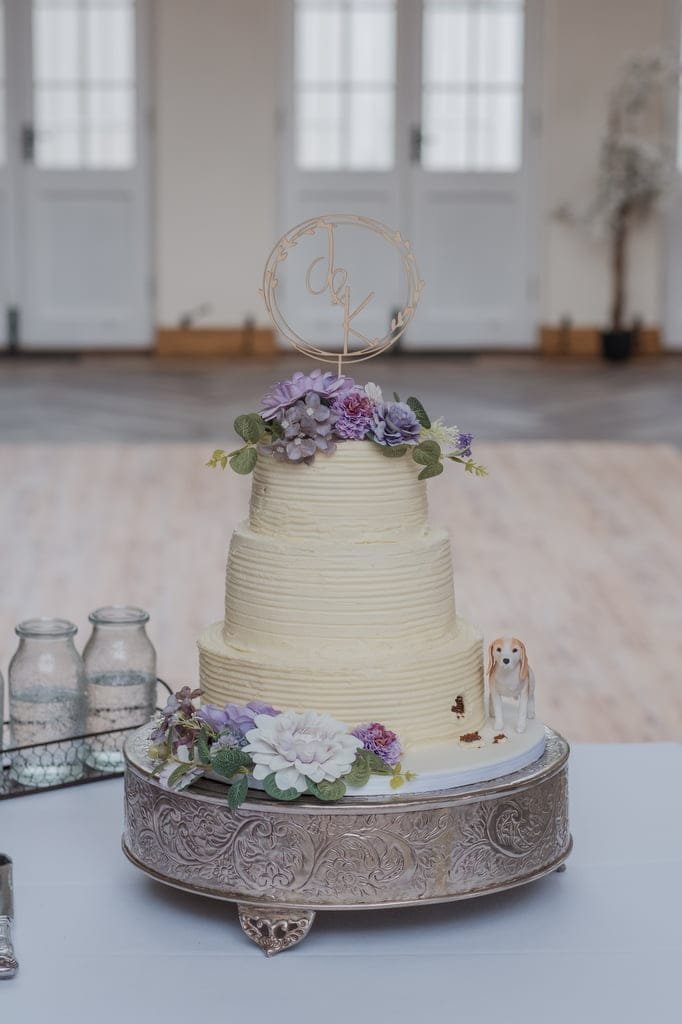 A three-tiered white wedding cake adorned with purple and white flowers, a figurine of a dog, and a wire cake topper sits on a silver cake stand, beautifully captured by your weddings photographer at Elmhay Park.
