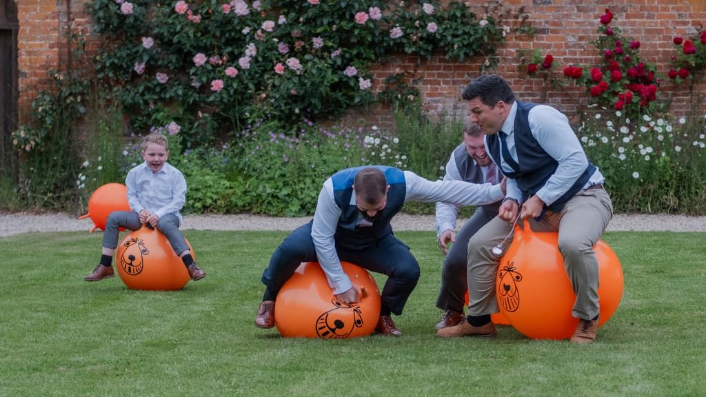 Four people, dressed in formal attire, are participating in a race on orange space hoppers in a garden with blooming flowers and a brick wall at the picturesque Elmhay Park—perfect for any weddings photographer to capture unique moments.