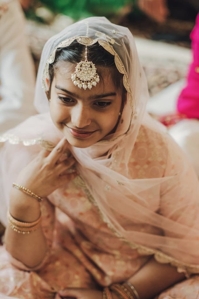 A young girl wearing a traditional pink outfit and headscarf adorned with a decorative headpiece and bangles, smiling gently, showcasing wedding traditions from around the world.