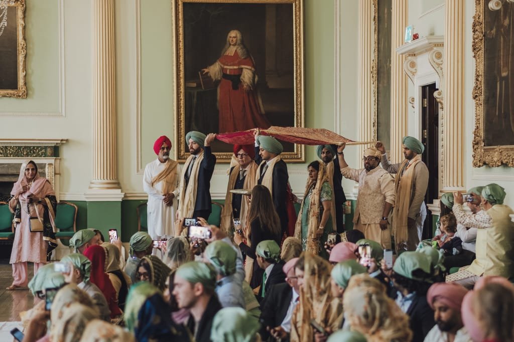 People dressed in traditional attire gather in the ornate Guildhall, with large portraits adorning the walls. Some hold umbrellas over those standing, while others sit, observing and capturing moments as a wedding photographer discreetly mingles among them.