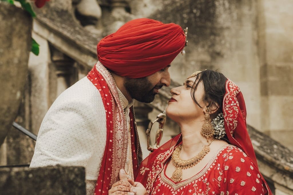 A couple in traditional red and white attire share an intimate moment, with the man wearing a turban and the woman adorned with jewelry, captured beautifully by Fuji Cameras for weddings, in front of stone steps.