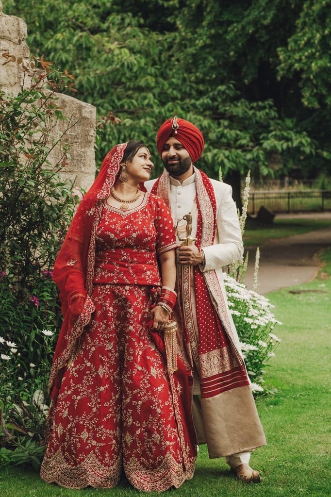 A couple dressed in traditional Indian wedding attire stands in a garden, embodying the vibrant Wedding Traditions from around the world. The woman wears a stunning red lehenga, while the man complements her with a white sherwani and a red turban.