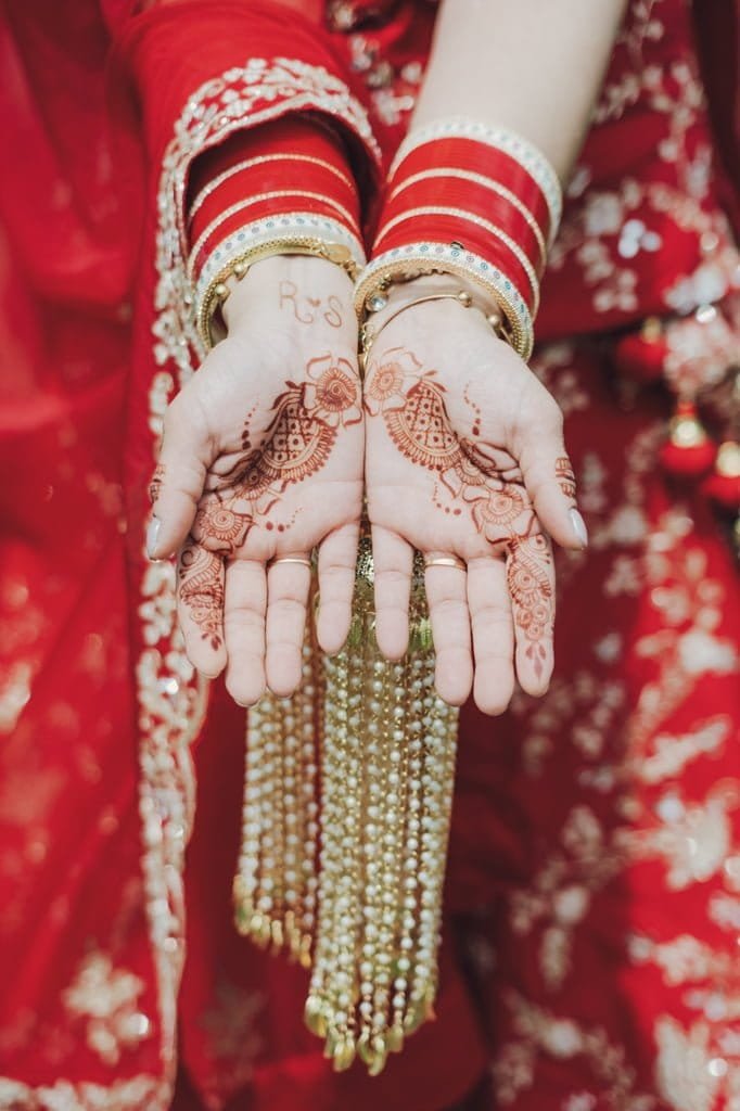 A person wearing red and gold bangles displays intricate mehndi designs on their open palms, with gold tassels hanging from their wrists, showcasing a beautiful aspect of wedding traditions from around the world.