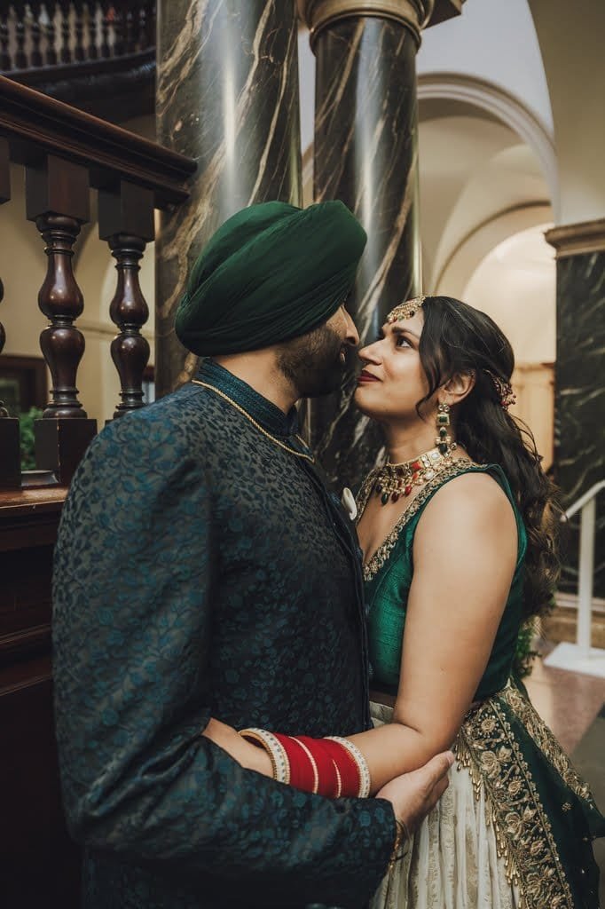 A couple dressed in traditional Indian attire embraces while gazing into each other's eyes inside a grand building, embodying wedding traditions from around the world. The man wears a green turban and ornate outfit; the woman is in a green and gold dress.