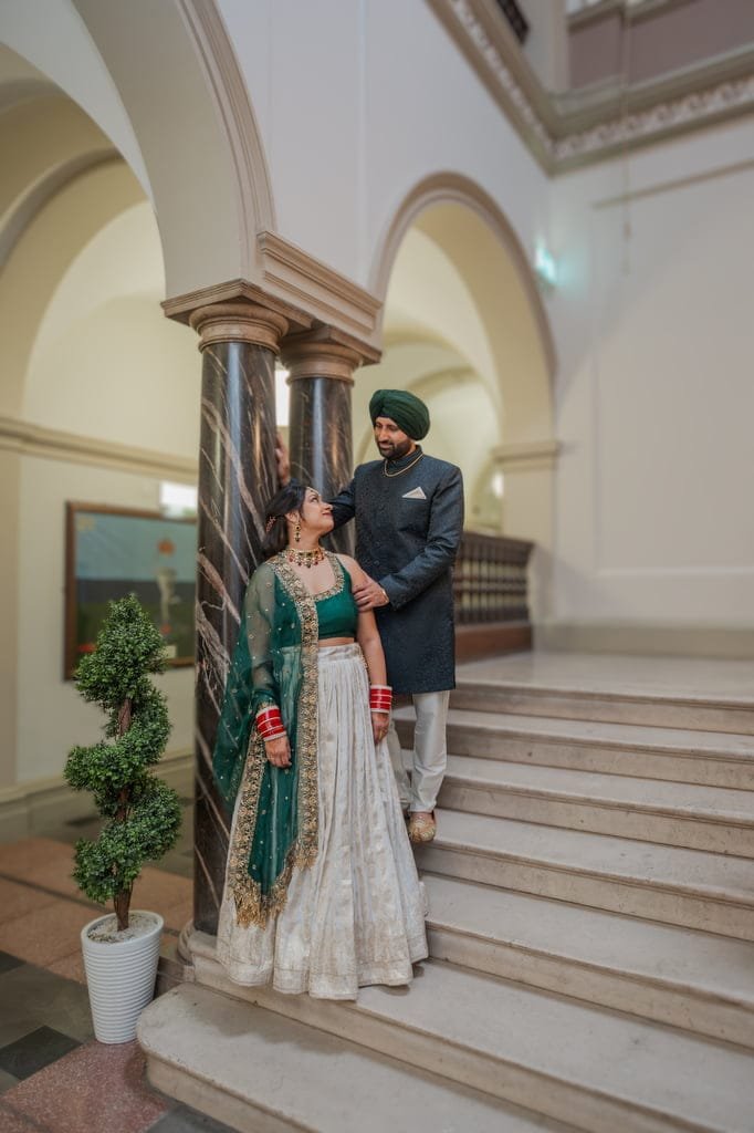 A couple dressed in traditional South Asian attire stands on an indoor staircase, looking at each other. The woman wears a green and gold outfit, and the man dons a dark-colored Sherwani with a green turban, captured beautifully by Guildhall Bath Wedding Photographer.