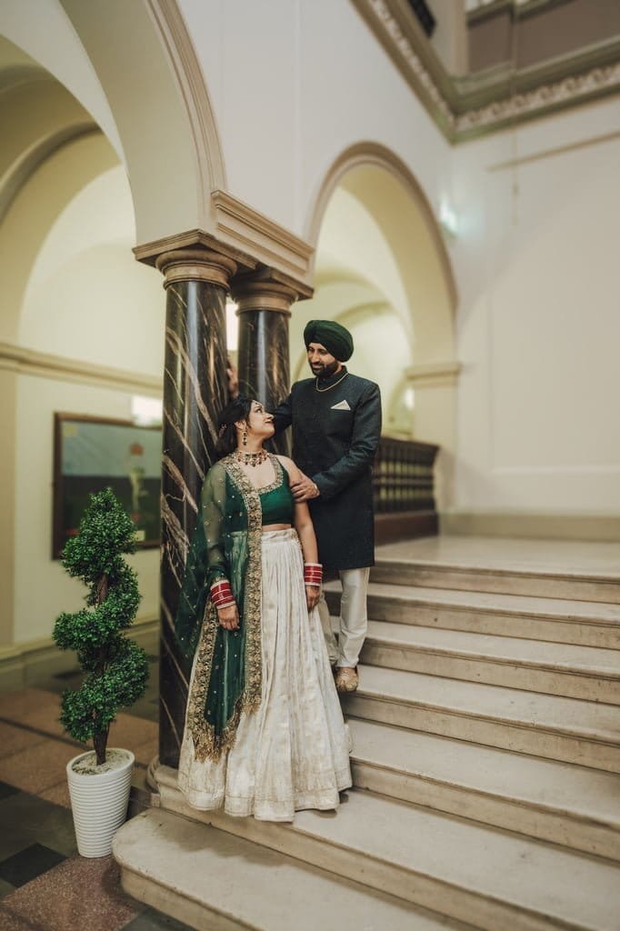 A couple stands on indoor stairs, embodying wedding traditions from around the world. The woman wears a green and white lehenga, while the man dons a dark green sherwani with a matching turban.