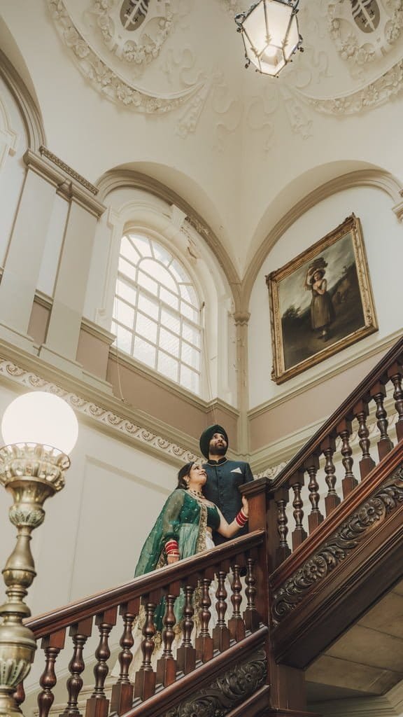 A couple stands on a staircase inside a building with ornate architecture; the woman wears traditional attire, and the man wears a turban and suit, reflecting wedding traditions from around the world. A painting and large window are visible in the background.