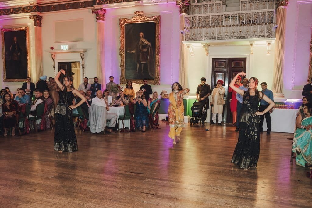 A group of performers in traditional attire dance inside an ornate hall, celebrating wedding traditions from around the world, with an audience seated at tables.