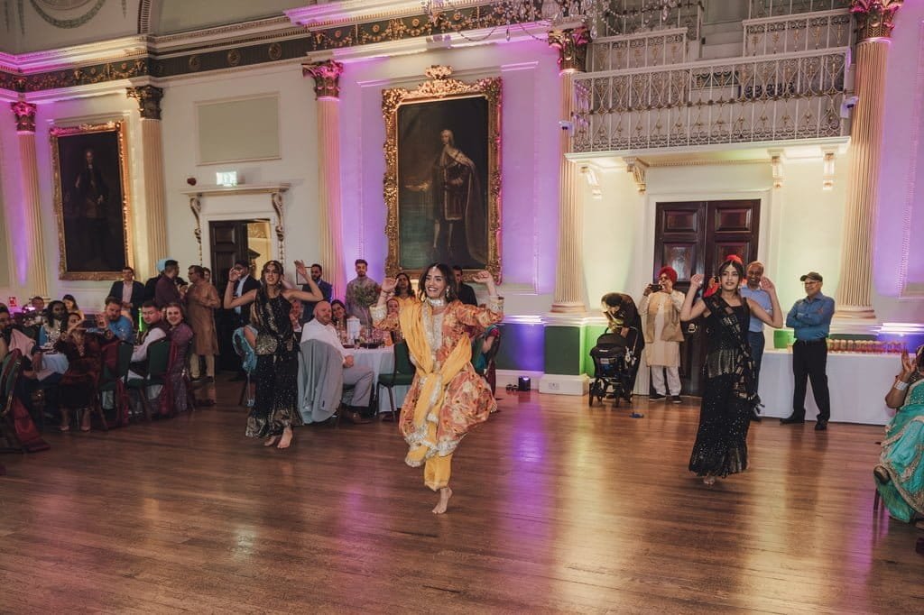 A group of people in a ballroom setting watches dancers in colorful attire perform, celebrating wedding traditions from around the world. The room is adorned with large paintings and ornate elements, adding to the festive atmosphere.