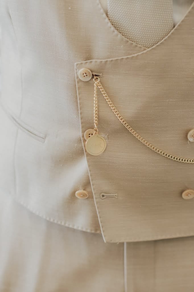 Close-up of a beige suit jacket with a matching vest and a gold pocket watch chain hanging from a button, captured beautifully by the Farleigh House Wedding Photographer.