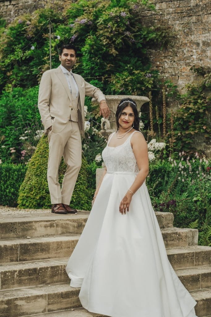 A couple in formal attire poses outdoors; the man stands on stone steps in a beige suit, and the woman, wearing a white gown, stands below him. Greenery and a stone wall serve as the backdrop, captured beautifully by our Farleigh House Wedding Photographer.