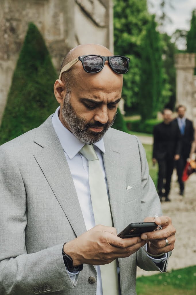 A man in a gray suit, white dress shirt, and light green tie is looking at his phone with sunglasses perched on his head. He stands outdoors with trees and people in the background, possibly scouting locations like a Farleigh House Wedding Photographer.