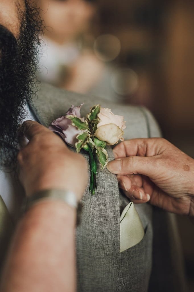 A Farleigh House Wedding Photographer captures a tender moment as a person pins a boutonnière with pink and lavender flowers onto the lapel of a man in a gray suit.