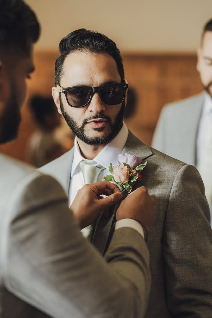 A man wearing sunglasses has a boutonnière adjusted on his suit jacket by another person. In the background, other people are visible, perhaps captured by a Farleigh House Wedding Photographer.