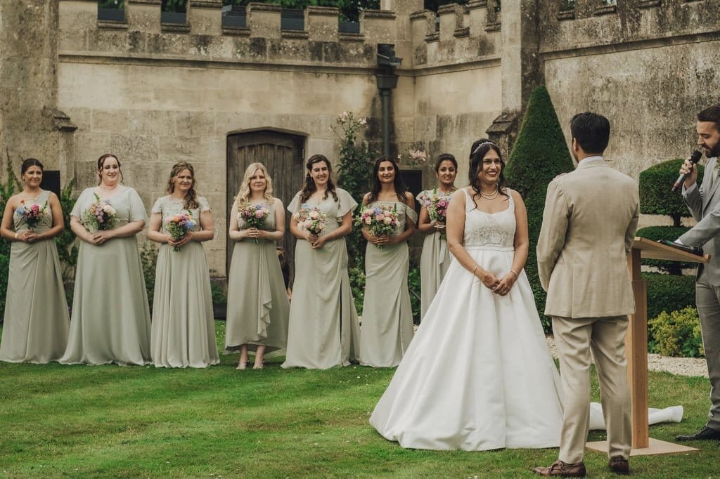 A Farleigh House wedding photographer captures the magic of a wedding ceremony outdoors, where a bride and groom stand before an officiant. A line of bridesmaids in light-colored dresses stands to the side, holding floral bouquets, perfectly framed by the scenic backdrop.