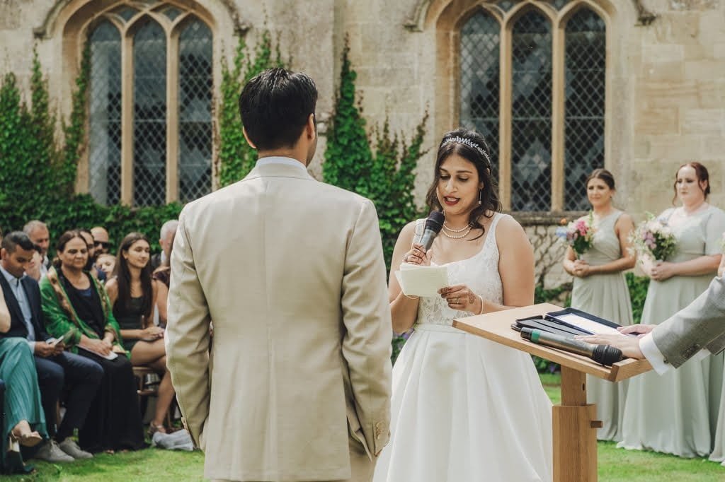 Bride holding a microphone and reading vows to the groom during an outdoor ceremony at Farleigh House, with bridesmaids and guests seated in the background.