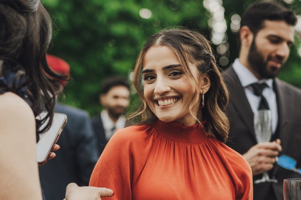 A woman in an orange top smiles at the camera at an outdoor gathering, with other attendees in the background, captured beautifully by a Farleigh House Wedding Photographer.