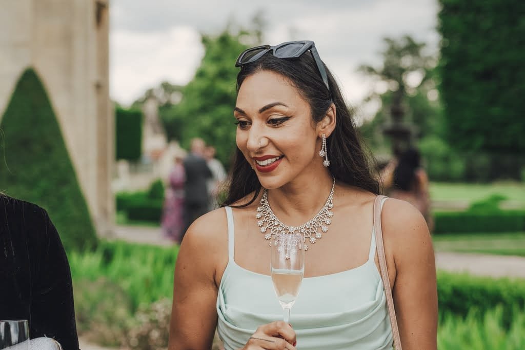 A woman in a light dress and sunglasses on her head holds a glass of champagne at what appears to be a Farleigh House wedding. She is wearing a statement necklace and earrings, with the lush garden setting providing the perfect backdrop for this elegant moment.