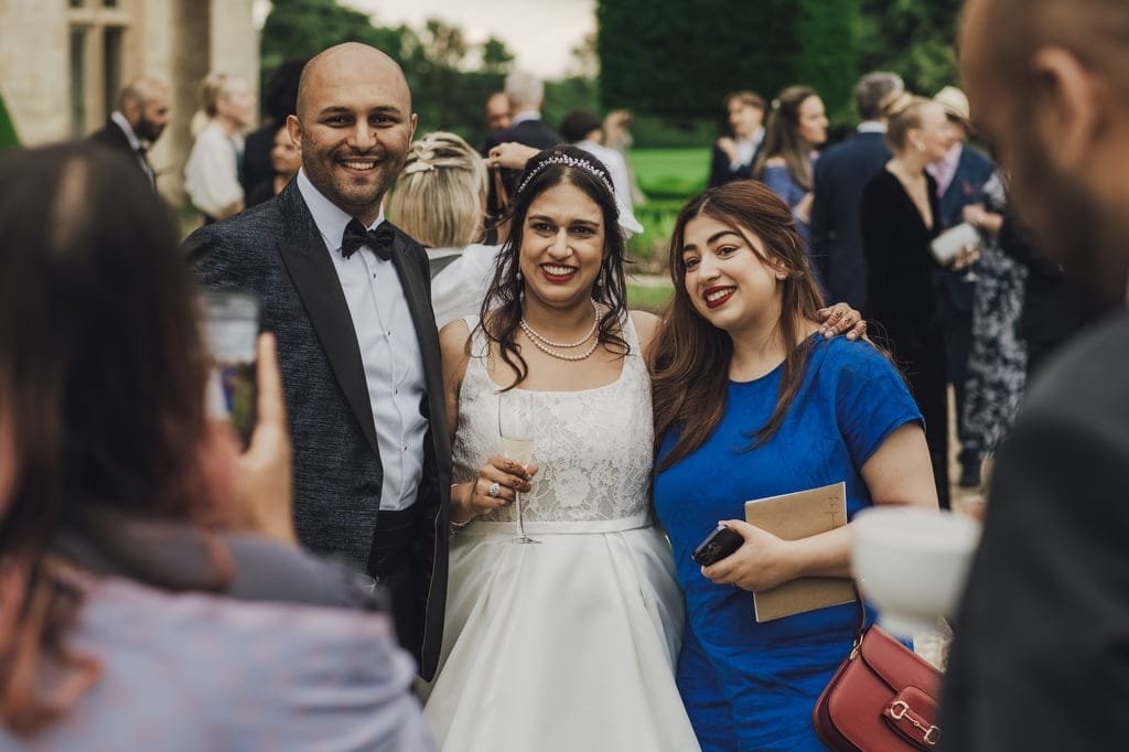 A bride in a white dress and a groom in a suit pose for a photo with a guest in blue attire at an outdoor wedding reception attended by others, captured beautifully by the Farleigh House Wedding Photographer.