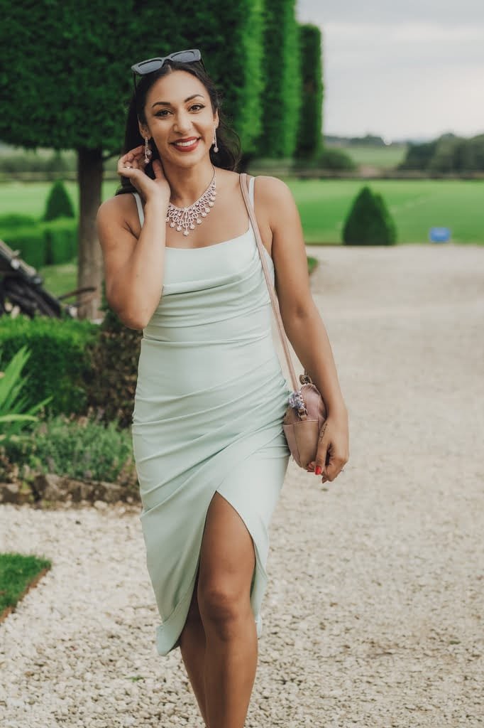 A woman in a light green dress and sunglasses on her head walks on a gravel path, smiling as she holds a brown handbag. Bushes and trees frame the scene, making it feel like it was captured by a Farleigh House Wedding Photographer.