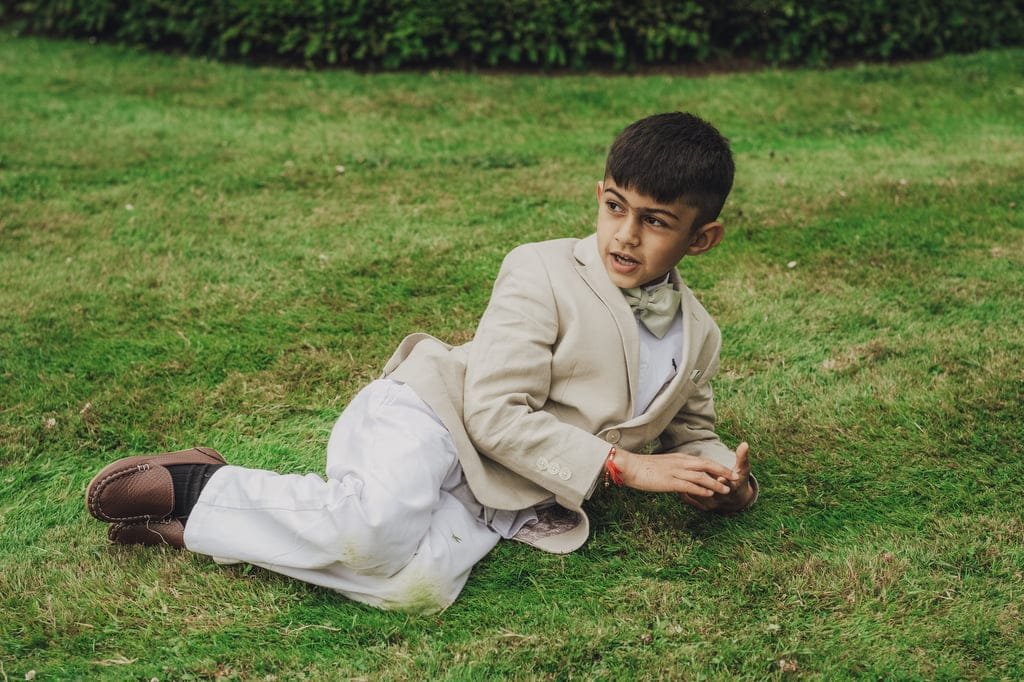 A young boy, dressed in a beige suit and bow tie, lies on a grassy lawn at Farleigh House. He gazes to his left with a curious expression, perfectly captured by the wedding photographer.