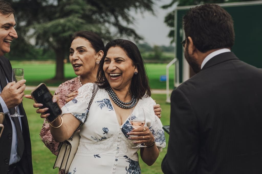 A group of four people stand outdoors, smiling and talking. One woman is holding a phone, while another woman is holding a glass of wine. Two men in suits are engaged in the conversation, perfectly captured by the Farleigh House Wedding Photographer.