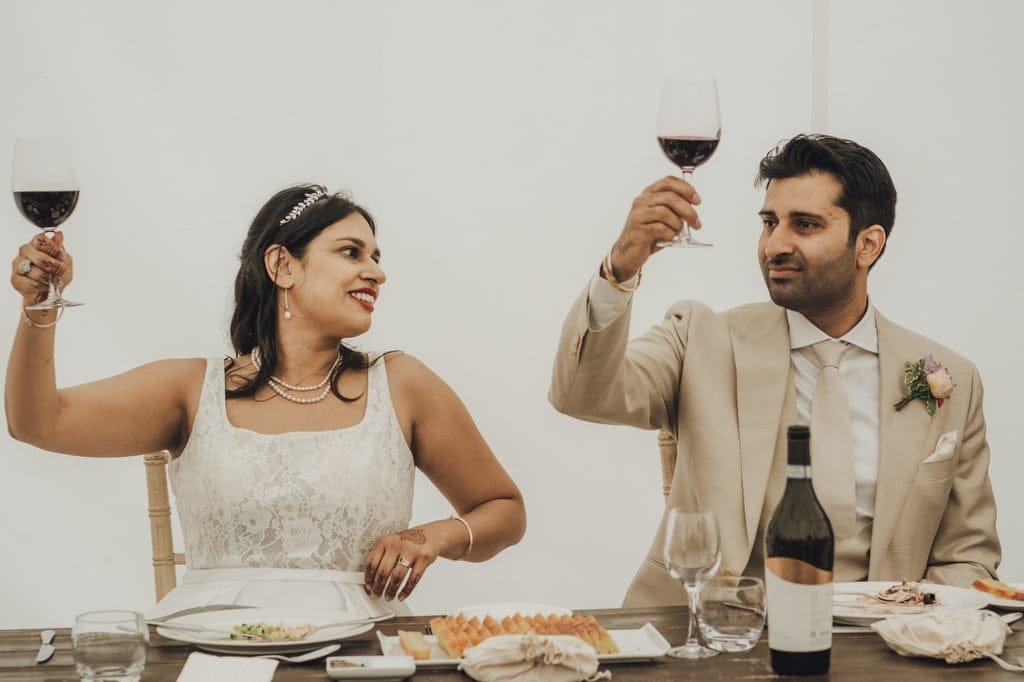 A bride and groom seated at a table, both raising wine glasses in a toast, with a bottle of wine and plates of food on the table in front of them, beautifully captured by a Farleigh House Wedding Photographer.